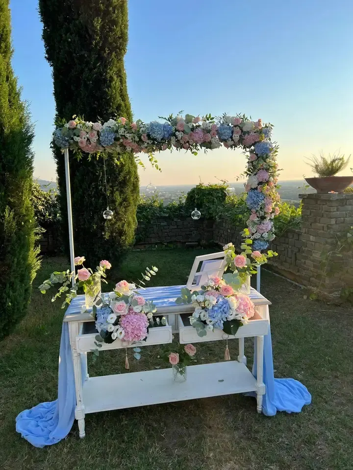 Tavolo cerimonia per battesimo: arco floreale con ortensie rosa e azzurre, candele sospese, tovaglie chiare, vista del tramonto sullo sfondo.