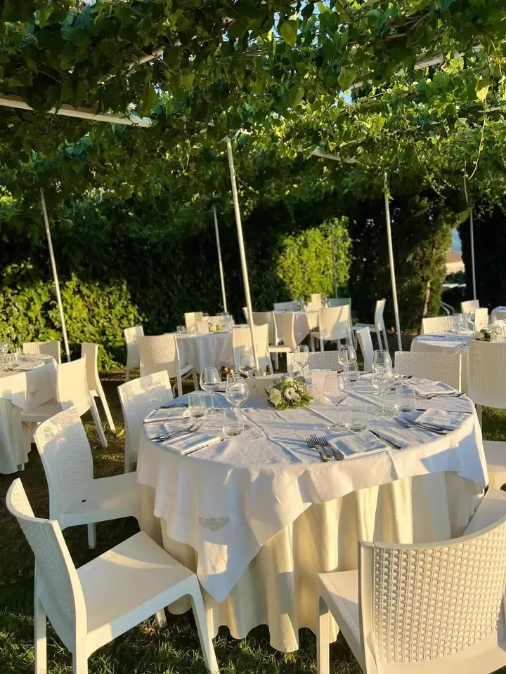 Tavolo rotondo apparecchiato sotto pergolato di vite: tovaglia bianca lunga, calici, fiori centrotavola, sedie bianche.