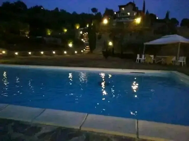 Piscina di sera con riflessi di luce sull'acqua, scalinata illuminata che sale verso la casa in cima alla collina, ombrellone e tavolini sul bordo.
