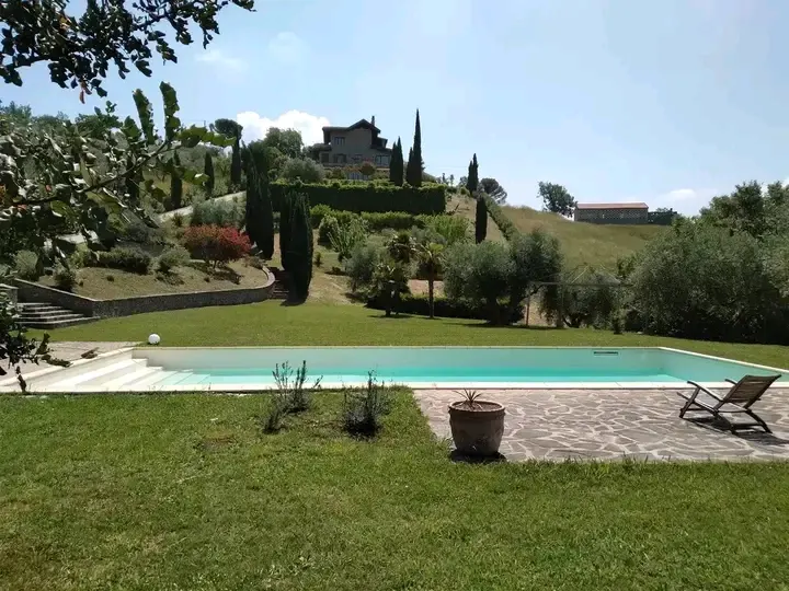 Piscina rettangolare con vista sulla collina: l'acqua riflette il cielo, sullo sfondo cipressi, ulivi, casa in pietra e vegetazione mediterranea.