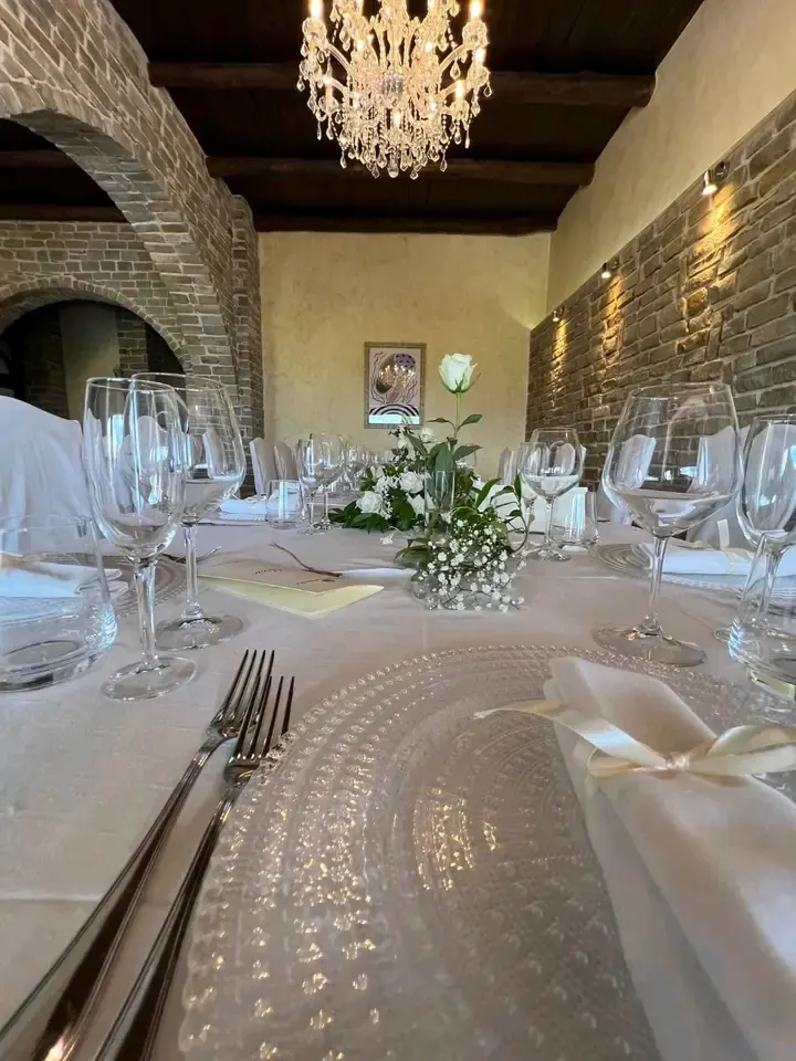 Sala interna per matrimonio: arco in pietra e mattoni a vista, lampadario di cristallo, soffitto con travi in legno antico, tavolo con calici, sottopiatto perlato e bouquet bianco.