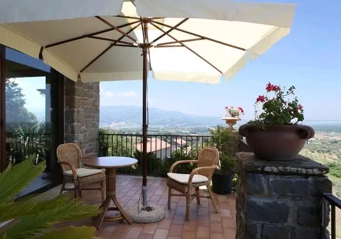 Terrazza con pavimento in cotto, tavolino bianco con due sedie in vimini, ombrellone color paglia, vaso con fiori rosa, vista panoramica.