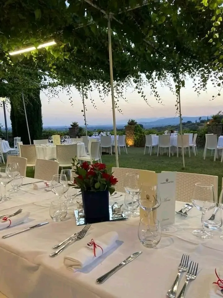 Tavoli allestiti sotto pergolato di vite al tramonto: bicchieri, segnaposto, menù Torre in Pietra in primo piano, vista delle colline in lontananza.
