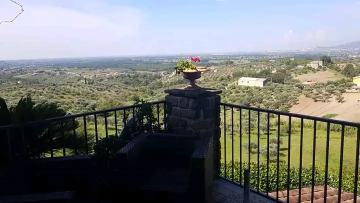 Vista dalla terrazza: vaso terracotta in primo piano, ringhiera in ferro, panorama di colline coltivate fino alla pianura.